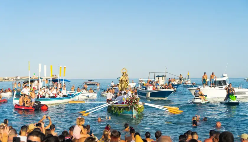 Procesión marítima con la Virgen del Carmen en un barco, rodeada de embarcaciones y gente en el agua, celebrando las fiestas.