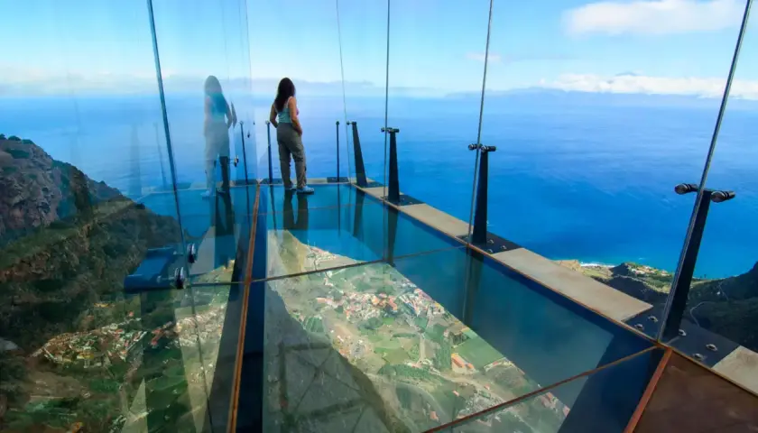 Mujer en mirador de cristal, 3 metros sobre el cielo, con vistas al mar y un pueblo.