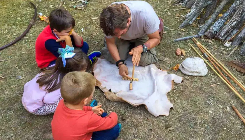 Un hombre enseña a niños a hacer fuego con un arco y un trozo de cuero. ¡Una aventura en la naturaleza para los más pequeños!