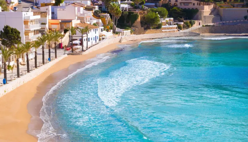 La playa de cala el Portet, con sus aguas turquesas y arena dorada, se extiende bajo un cielo azul, flanqueada por palmeras y casas blancas.