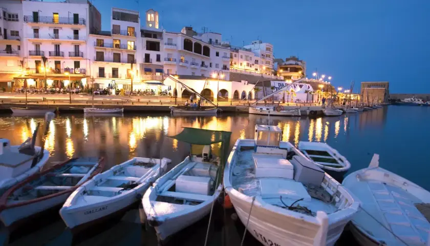 Noche en el paseo marítimo de L'Ametlla de Mar. Barcas blancas flotan en el puerto, reflejando las luces de los edificios y restaurantes.