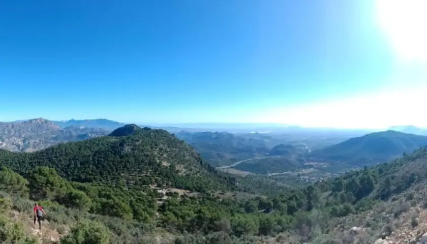Un excursionista disfruta de la vista panorámica desde el balcon de Alicante, un paisaje montañoso cubierto de pinos bajo un cielo azul intenso.