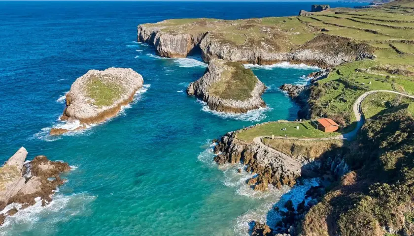 Acantilados y aguas turquesas en la costa de Cantabria. Un lugar idílico que ver en Isla Cantabria, con islotes rocosos y un sendero que lleva a una cabaña.
