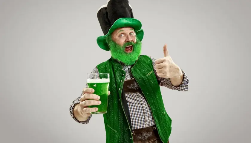 Hombre con barba verde y sombrero de duende, sosteniendo una cerveza verde y dando pulgar arriba. ¡Listo para el desfile de San Patricio!