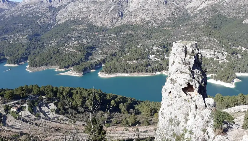 Impresionante vista del embalse de Guadalest con su agua turquesa, rodeado de montañas y pinos. Una roca solitaria se alza como centinela.