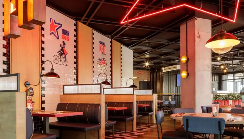 Interior de restaurante con cabinas, mesas y decoración moderna. Las paredes tienen murales inspiradores, como "Think Big". Las luces rojas de neón añaden un toque vibrante. Ideal para leer reseñas de Foster's Hollywood.