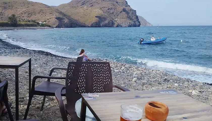 Una persona contempla el mar en la playa de las negras opiniones. Un bote azul descansa en el agua, mientras las olas acarician la costa pedregosa.