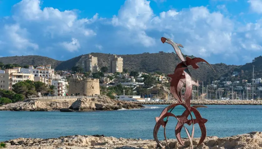 Escultura de aves sobre rocas, con el pueblo de Moraira y su torre defensiva al fondo.