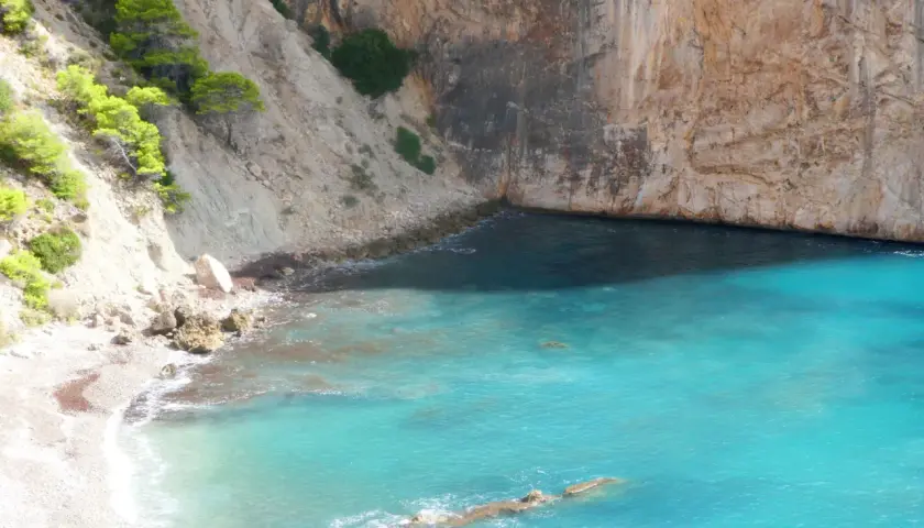 Aguas turquesas bañan la cala racó del corb, rodeada de imponentes acantilados y vegetación verde.