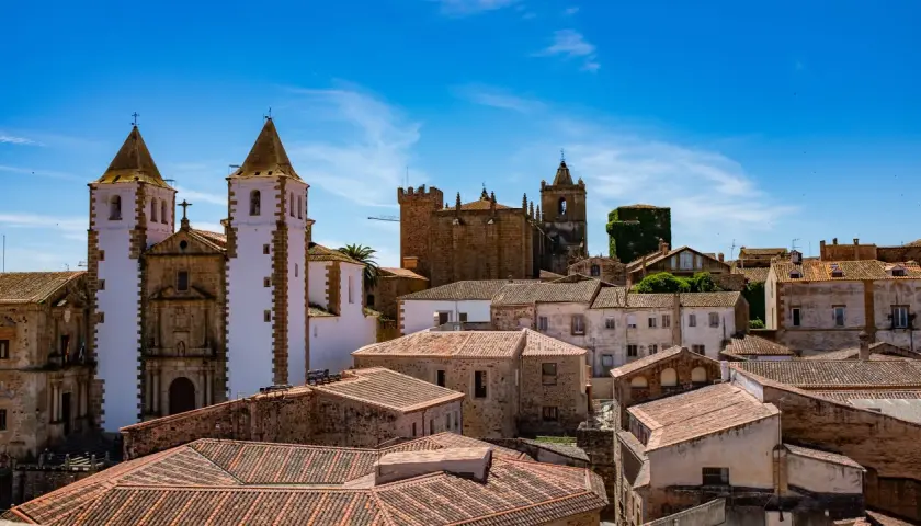 Vistas de tejados y edificios históricos, incluyendo iglesias con torres blancas y campanarios, bajo un cielo azul. Un lugar ideal que ver en Cáceres provincia.