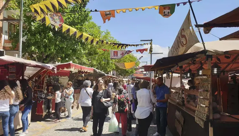 Gente pasea por el mercadillo medieval El Álamo, con puestos de artesanía y comida bajo banderines coloridos.