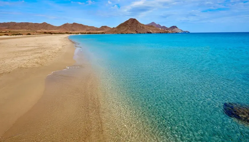 Arena dorada y aguas cristalinas en una de las playas bonitas en España, con montañas áridas al fondo bajo un cielo azul.