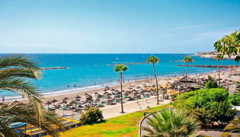 Un día soleado en el paseo marítimo de Playa de las Américas, con gente disfrutando de la playa, sombrillas y palmeras.
