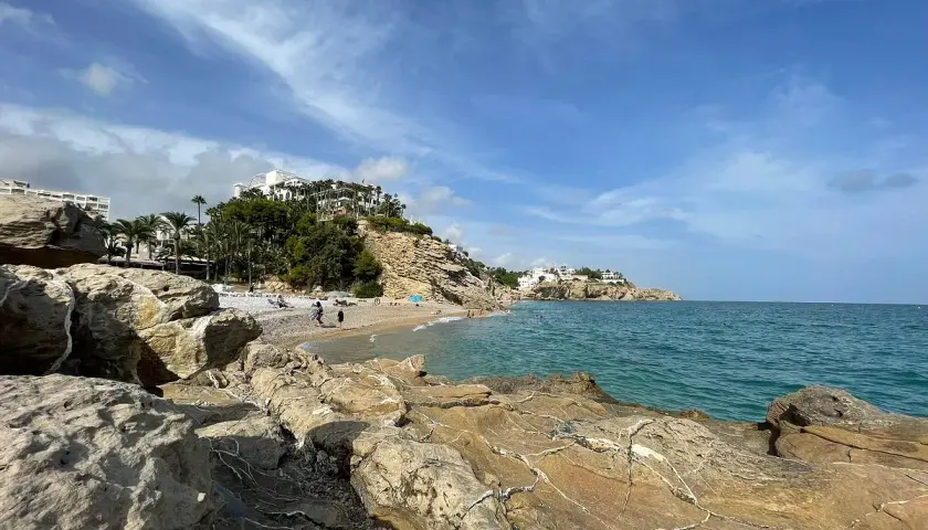Rocas doradas en primer plano, la caleta Villajoyosa con gente disfrutando del mar azul y edificios blancos en la colina.
