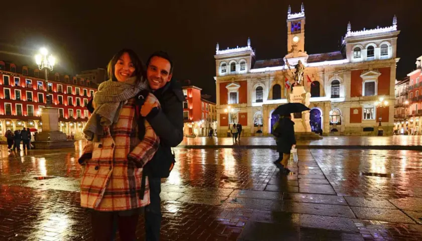 Pareja sonriente en la Plaza Mayor de Valladolid, un lugar bonito que ver en Valladolid, con edificios iluminados y el suelo mojado por la lluvia.
