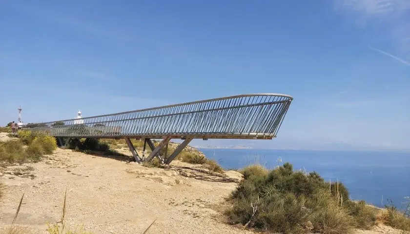 El mirador del faro de Santa Pola se proyecta sobre el mar azul, con vegetación árida y un cielo despejado.