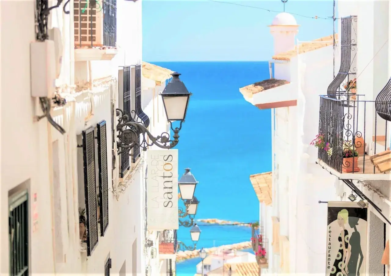 Calle empedrada con vistas al mar azul. Farolas y balcones floridos invitan a descubrir las cosas que hacer en Altea.