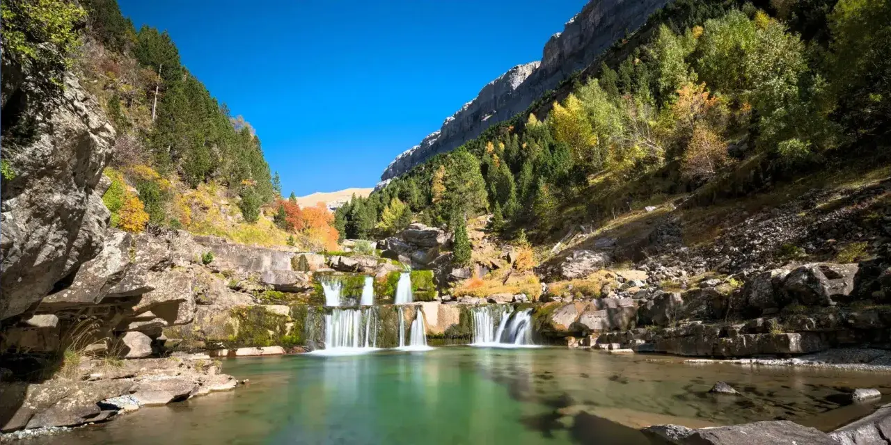 Cascada en un valle montañoso, ideal para rutas senderismo España. El agua verde turquesa fluye entre rocas y árboles otoñales.