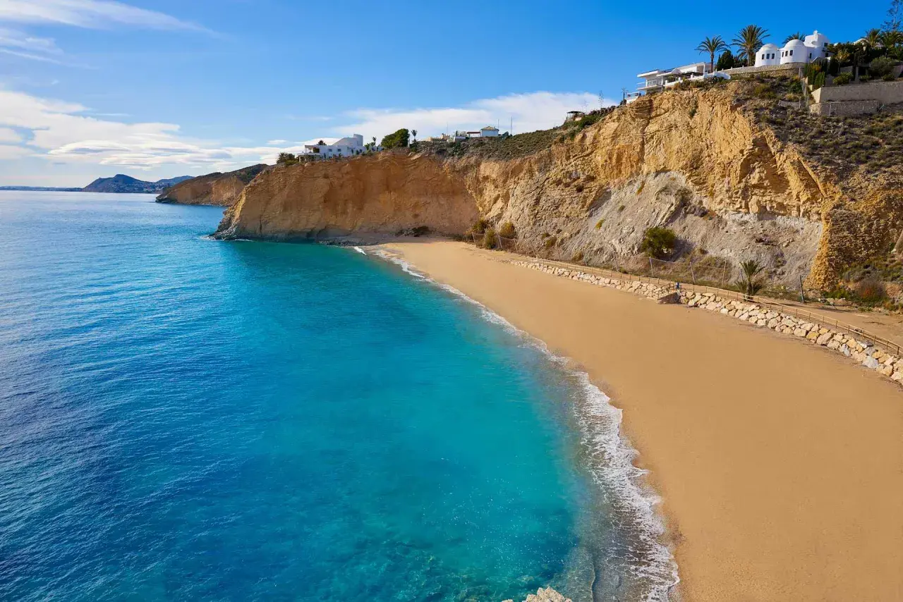 Arena dorada y aguas turquesas en la playa de Villajoyosa, con acantilados y casas blancas al fondo.