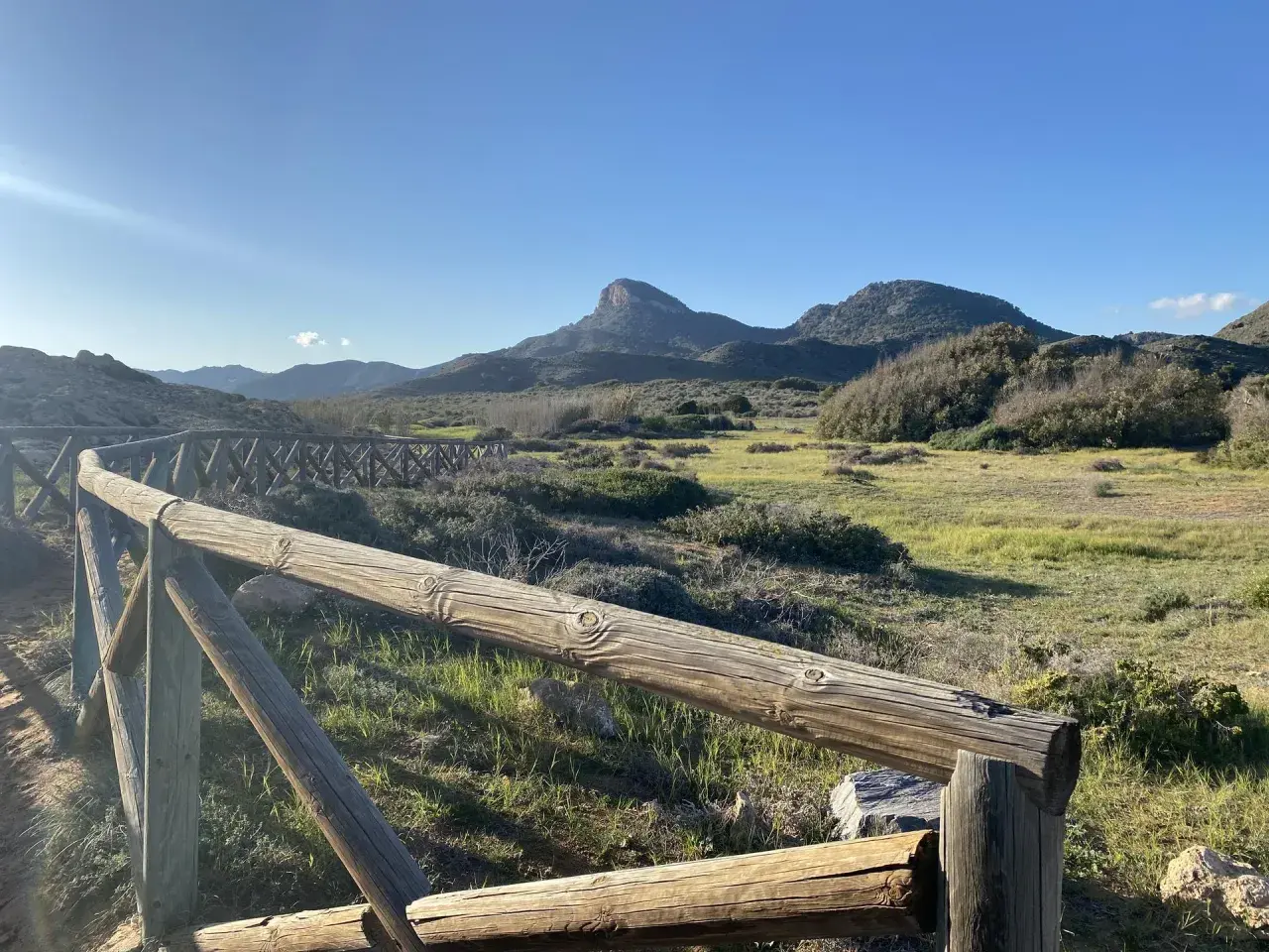 Un sendero de madera bordea un paisaje verde y montañoso, con el imponente monte de las cenizas al fondo bajo un cielo azul.