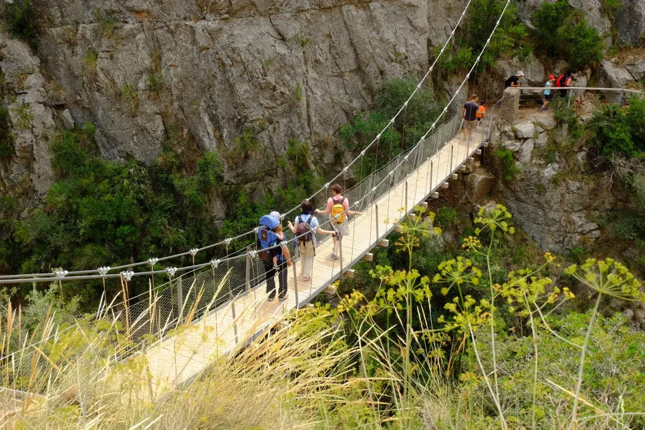 Senderismo en las **rutas Comunidad Valenciana**. Personas cruzan un puente colgante entre riscos y vegetación.
