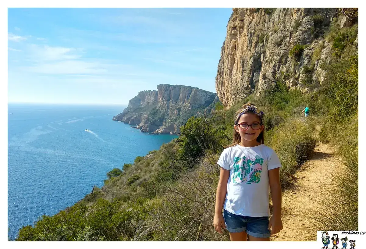 Niña sonriente en la ruta acantilados Benitachell, con el mar azul y formaciones rocosas al fondo.