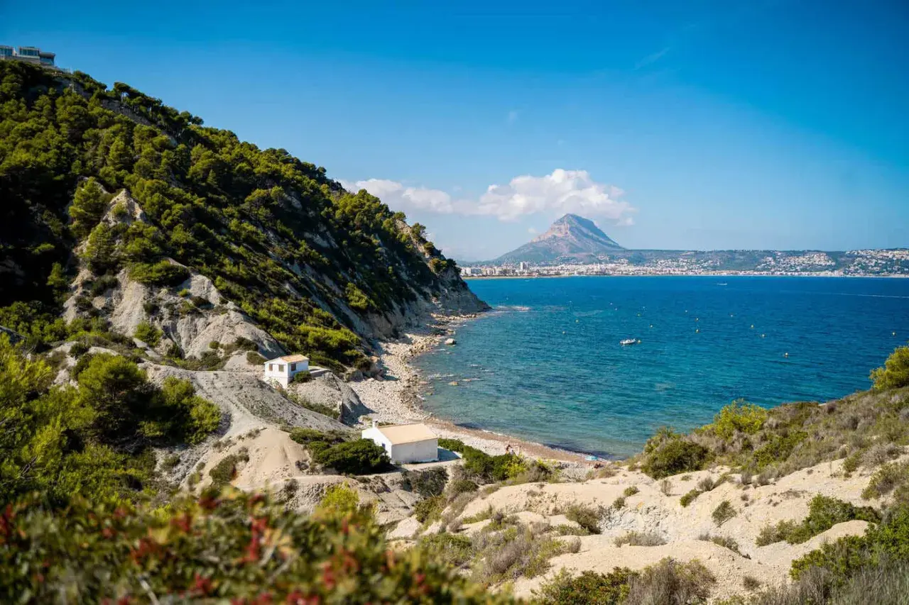 La cala del francés, con sus casitas blancas y el mar azul, se asoma a un paisaje rocoso y verde. Al fondo, la silueta de una montaña domina la costa.