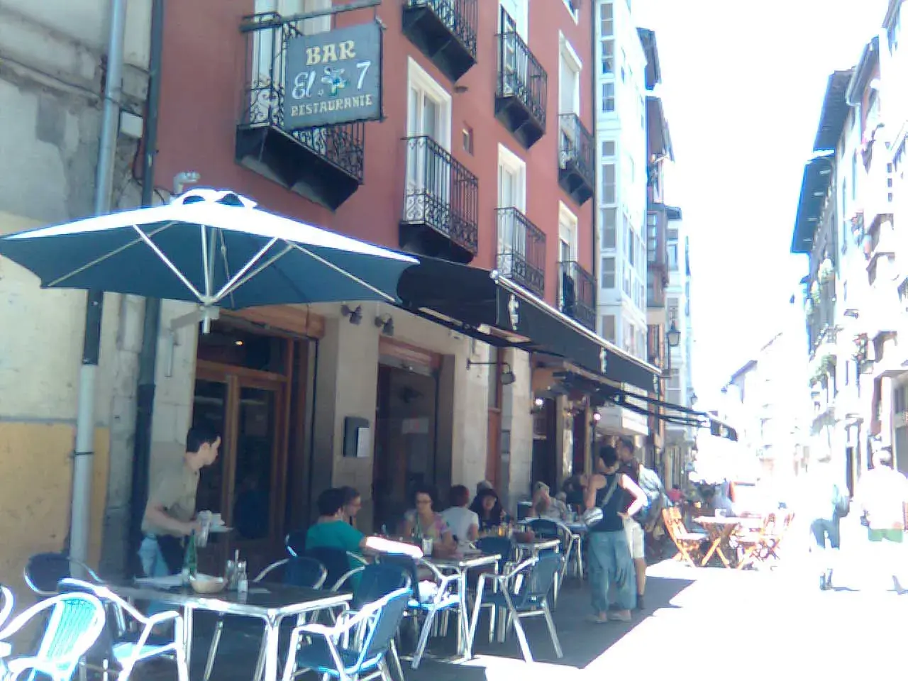 Gente disfrutando en las mesas exteriores del restaurante El 7 Vitoria, bajo un gran parasol azul.