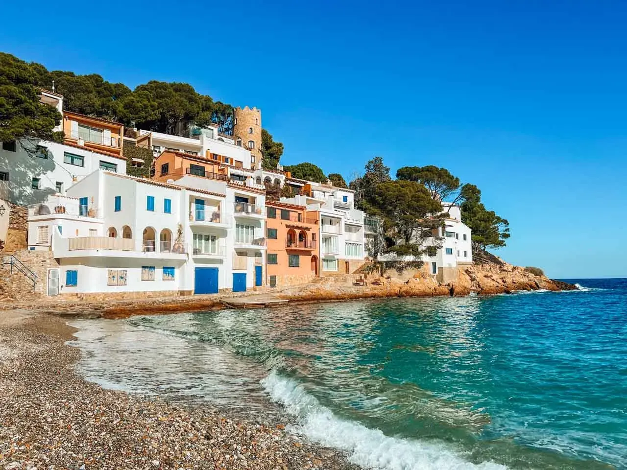 Casas blancas y coloridas se asoman al mar turquesa en la costa brava españa. Una pequeña playa de guijarros recibe las olas.