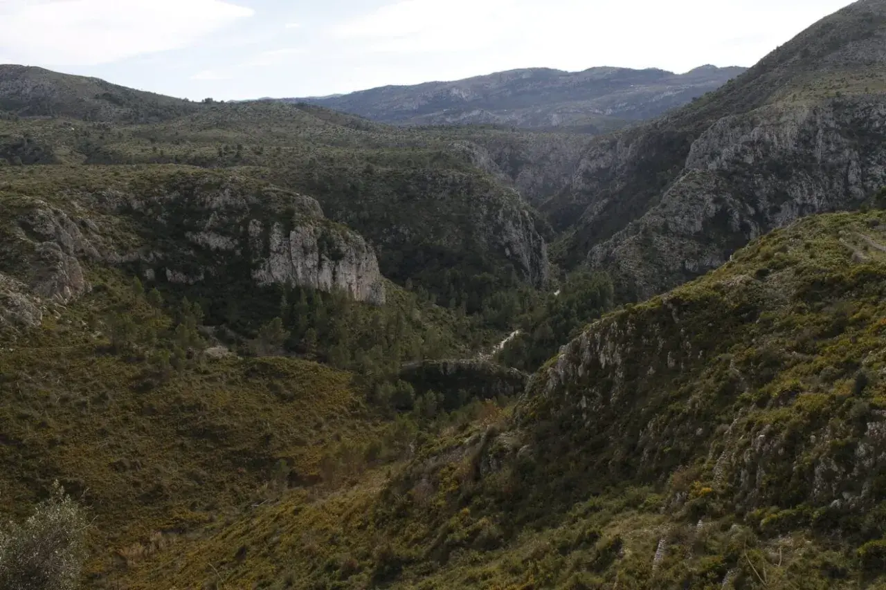 Paisaje montañoso con vegetación verde y rocas escarpadas. El **barranc de l'infern** se abre paso entre las colinas, con un camino serpenteante.