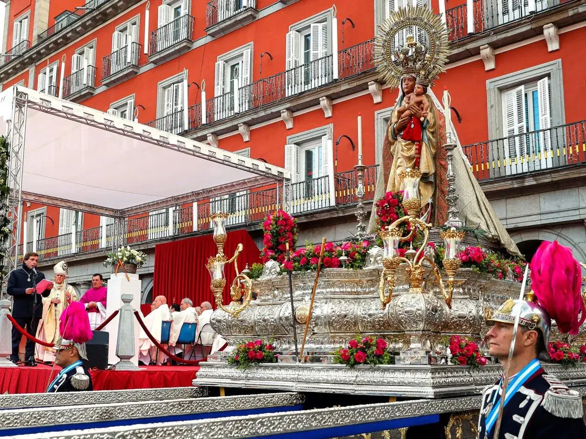 Procesión de la Almudena en Madrid. Una imagen de la Virgen con el Niño sobre un trono adornado con flores, con el fondo de edificios rojos y gente vestida de gala.