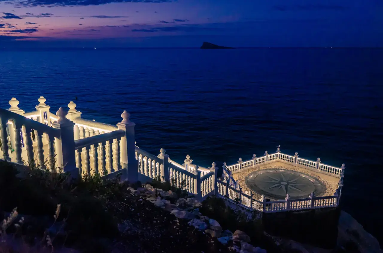 El mirador de Benidorm de noche, con el mar azul oscuro y un cielo crepuscular. Una balaustrada blanca ilumina el camino hacia una plataforma circular con una rosa de los vientos.