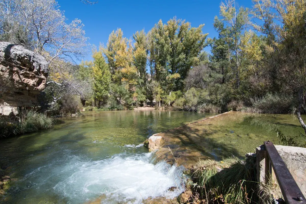 Un río de aguas cristalinas con un pequeño salto de agua. Al fondo, árboles con hojas otoñales y un cielo azul. ¡Un lugar con **alto tajo que ver**!