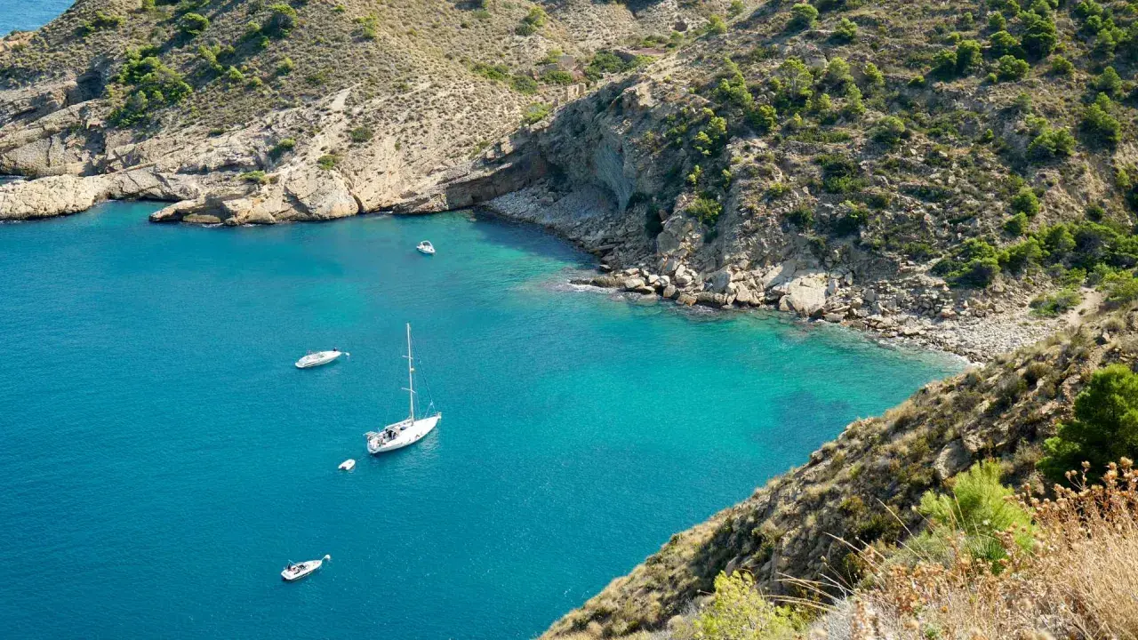 La cala del metge, con aguas turquesas y barcos navegando, se abre paso entre acantilados rocosos y vegetación mediterránea.