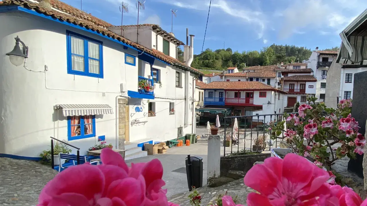 Casas blancas con detalles azules, tejados rojos y flores rosadas en un día soleado. Un vistazo a los encantadores pueblos cerca de Cudillero.