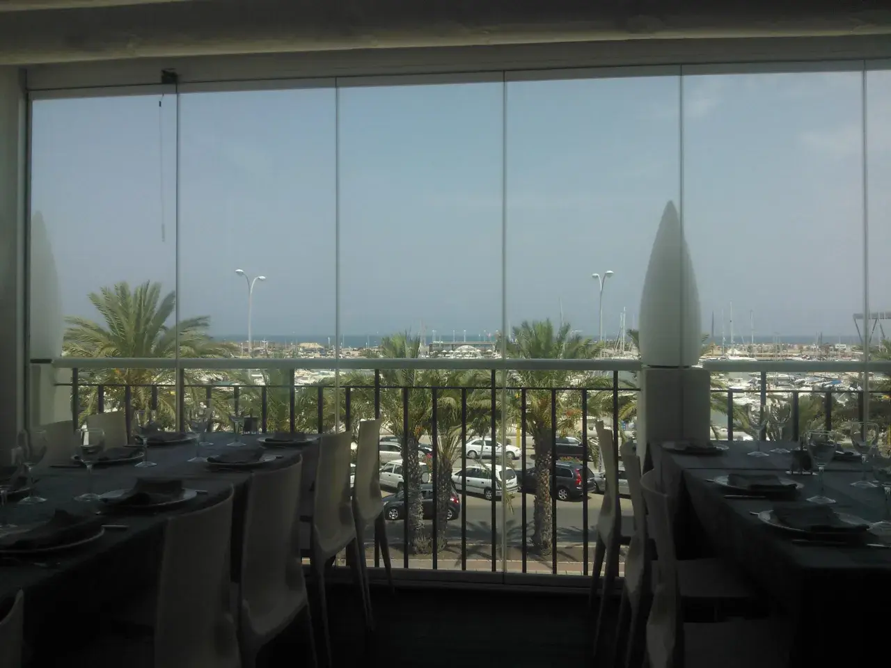 Mesas puestas con vistas al puerto deportivo de la barqueta Denia. Palmeras y barcos crean un ambiente vacacional.