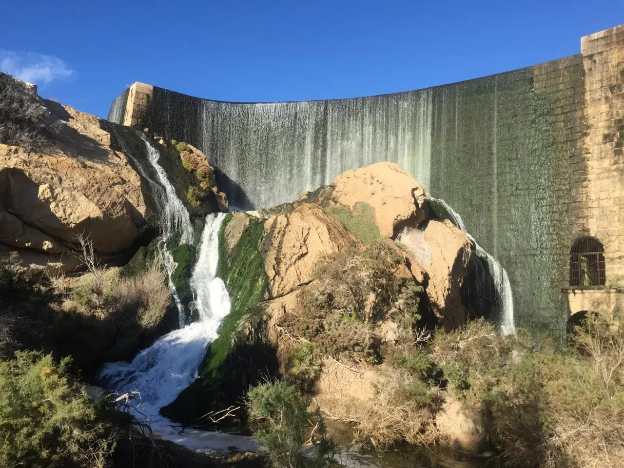 Cascada en la presa de Elche, un lugar ideal para una ruta pantano elche. El agua cae sobre rocas cubiertas de musgo y vegetación.