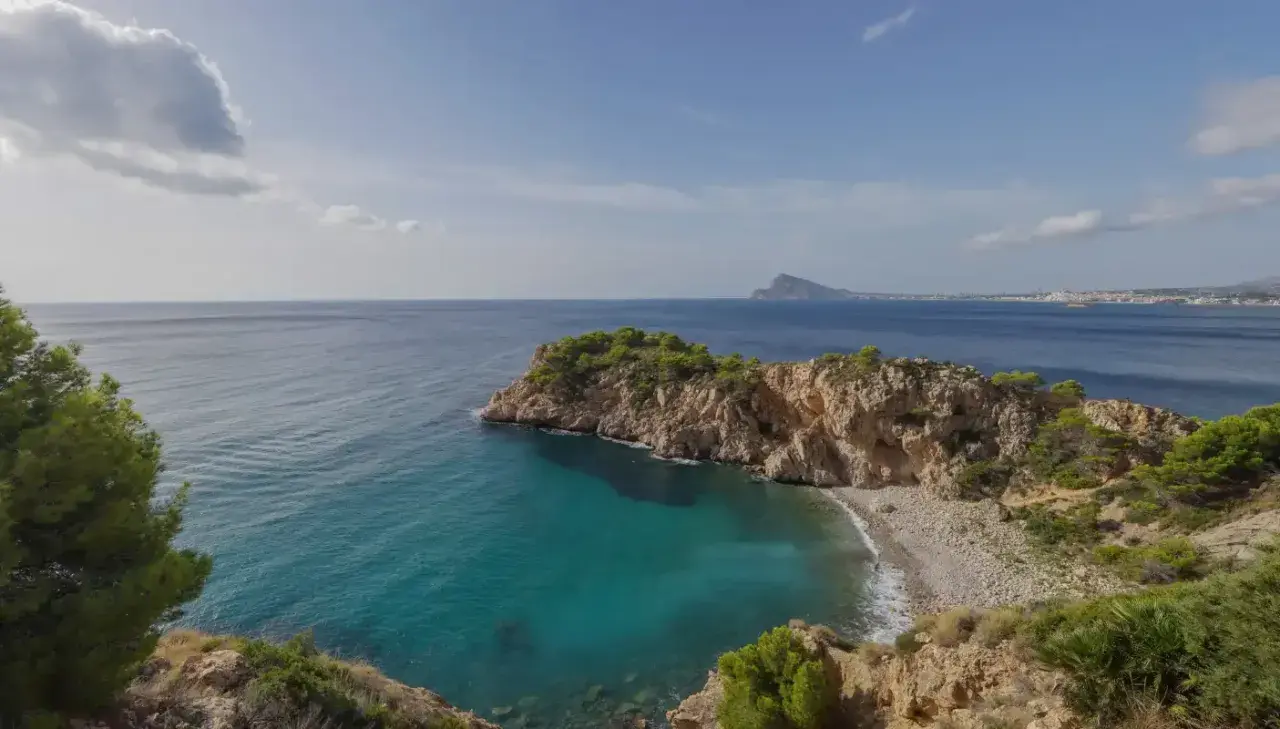 Vista de una cala con aguas turquesas y una gran barra de guijarros, rodeada de acantilados rocosos y vegetación.