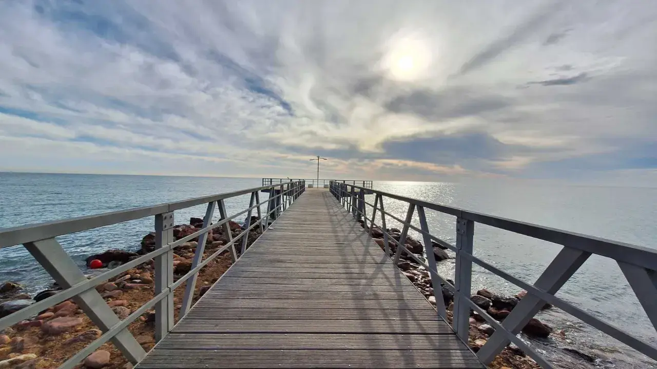 Muelle de madera sobre el mar, con cielo nublado y sol brillante. Un lugar ideal para disfrutar de la playa de Chilches.