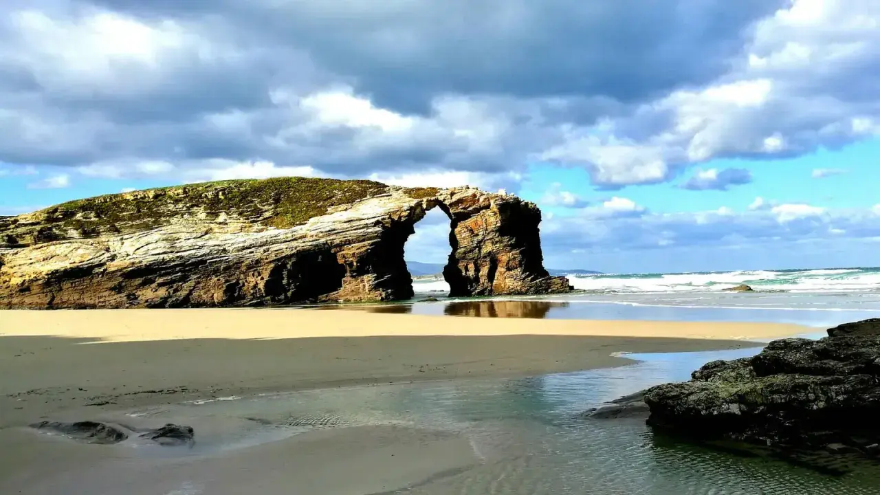 Arco natural en una playa de arena dorada, con el mar y nubes dramáticas. Un ejemplo de las mejores playas Galicia.