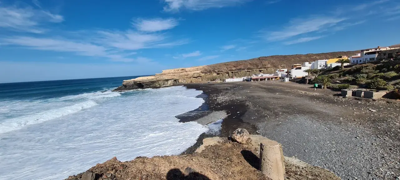 Olas rompen en la playa de arena negra de los charcones de Ajuy, con casas blancas al fondo y acantilados dorados.