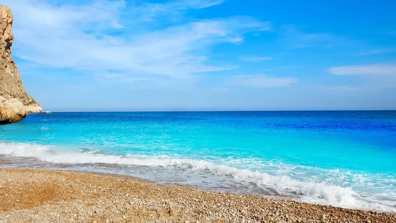 Aguas turquesas bañan la playa de guijarros de cala dels testos, con un acantilado rocoso a la izquierda y un cielo azul despejado.