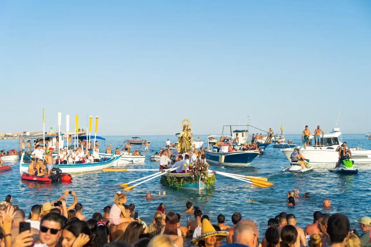 Procesión marítima con la Virgen del Carmen en un barco, rodeada de embarcaciones y gente en el agua, celebrando las fiestas.