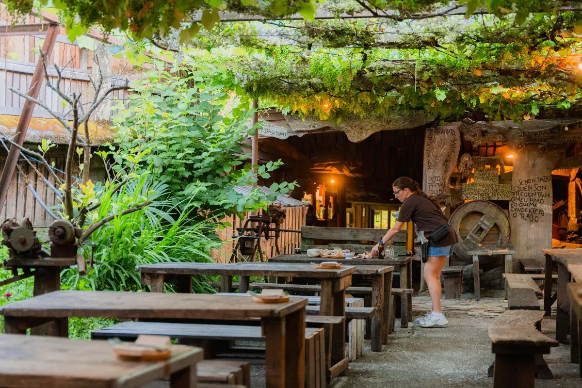 Terraza rústica del restaurante Fogar do Santiso, con mesas de madera bajo un toldo de hiedra. Una mujer prepara una mesa.