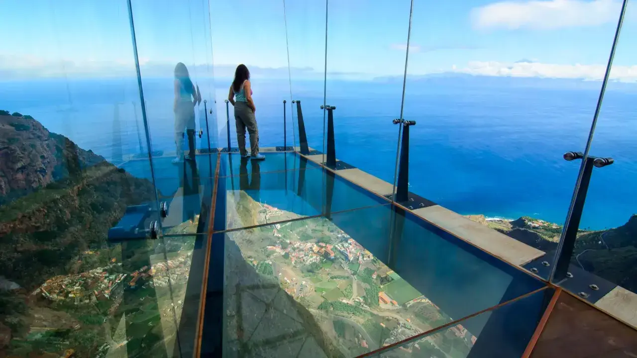 Mujer en mirador de cristal, 3 metros sobre el cielo, con vistas al mar y un pueblo.