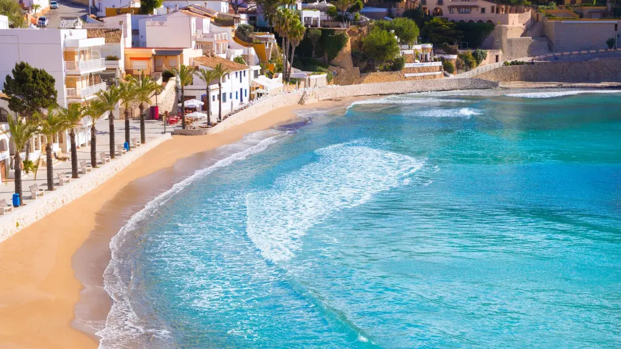 La playa de cala el Portet, con sus aguas turquesas y arena dorada, se extiende bajo un cielo azul, flanqueada por palmeras y casas blancas.