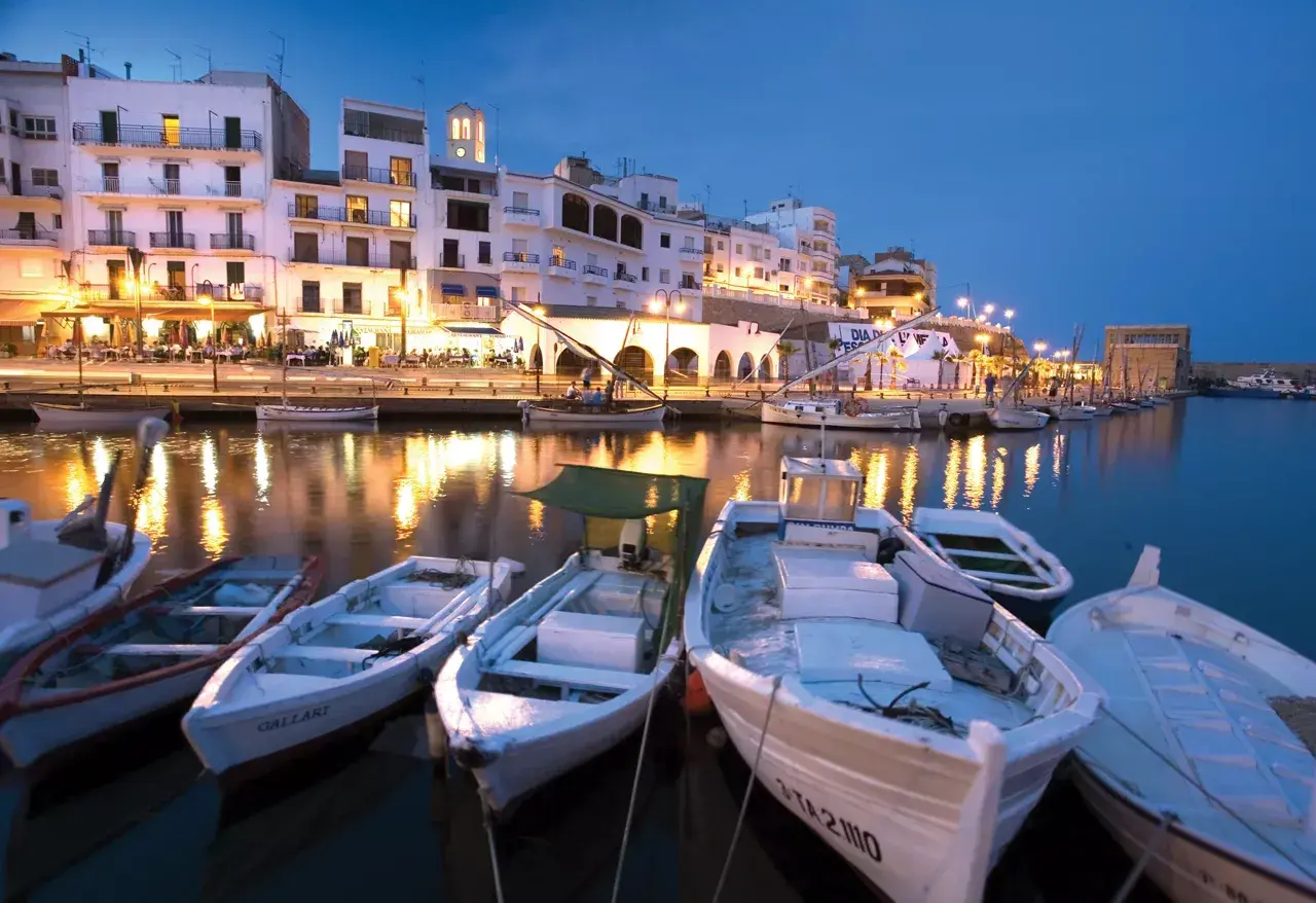 Noche en el paseo marítimo de L'Ametlla de Mar. Barcas blancas flotan en el puerto, reflejando las luces de los edificios y restaurantes.