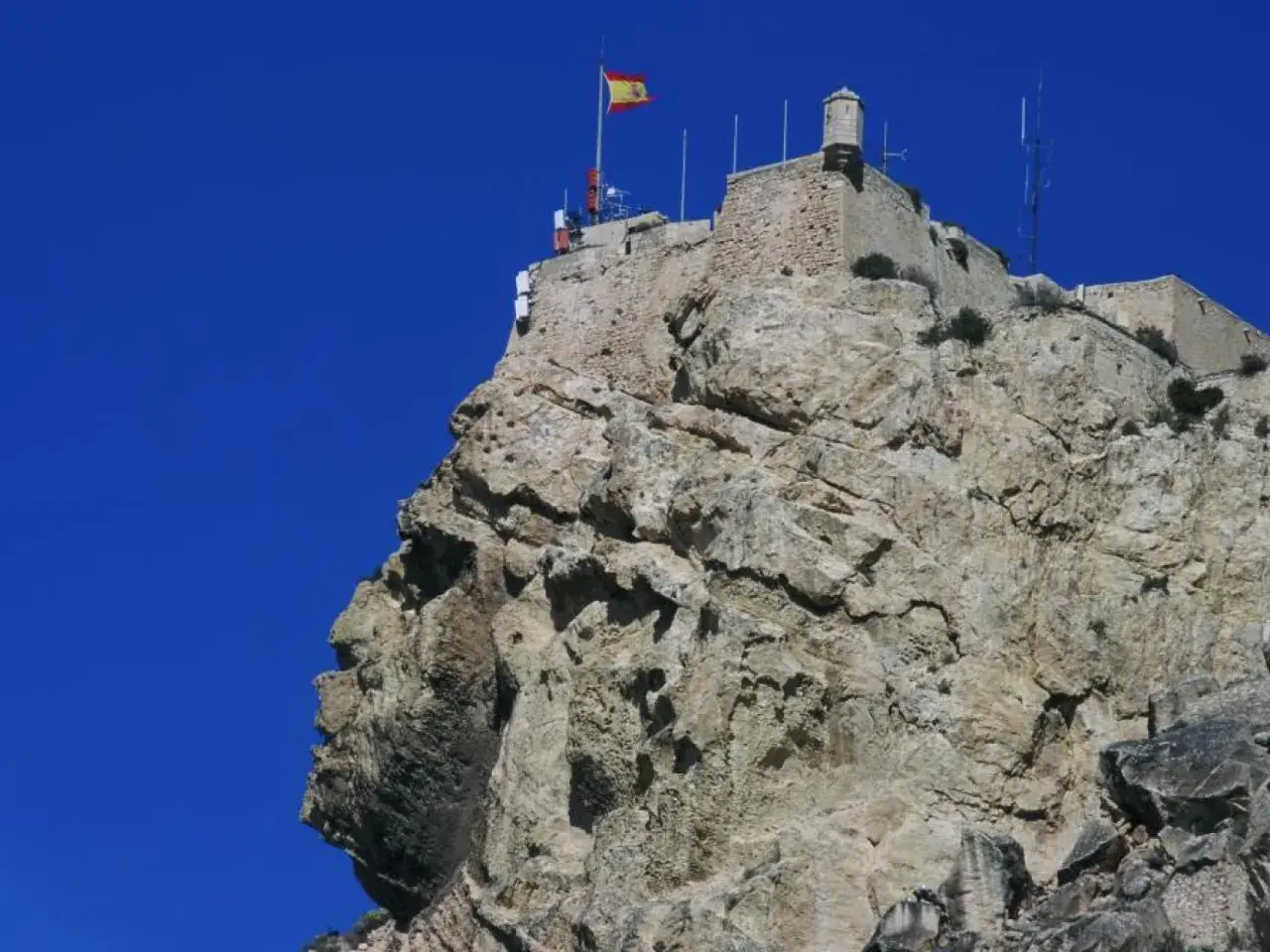 La cara del moro alicante, una roca con forma de perfil humano, corona la cima de una montaña rocosa con un castillo y una bandera española.