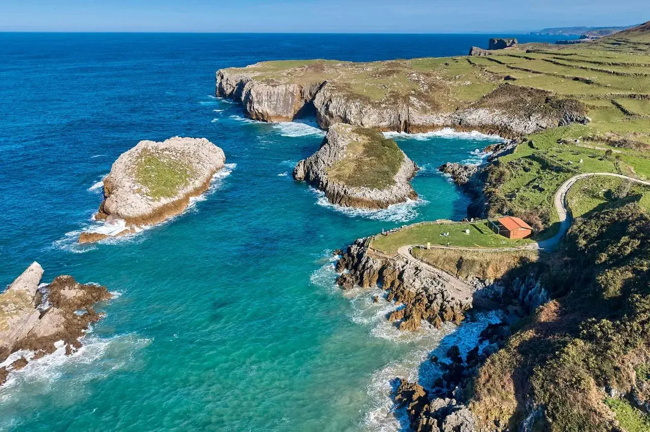 Acantilados y aguas turquesas en la costa de Cantabria. Un lugar idílico que ver en Isla Cantabria, con islotes rocosos y un sendero que lleva a una cabaña.
