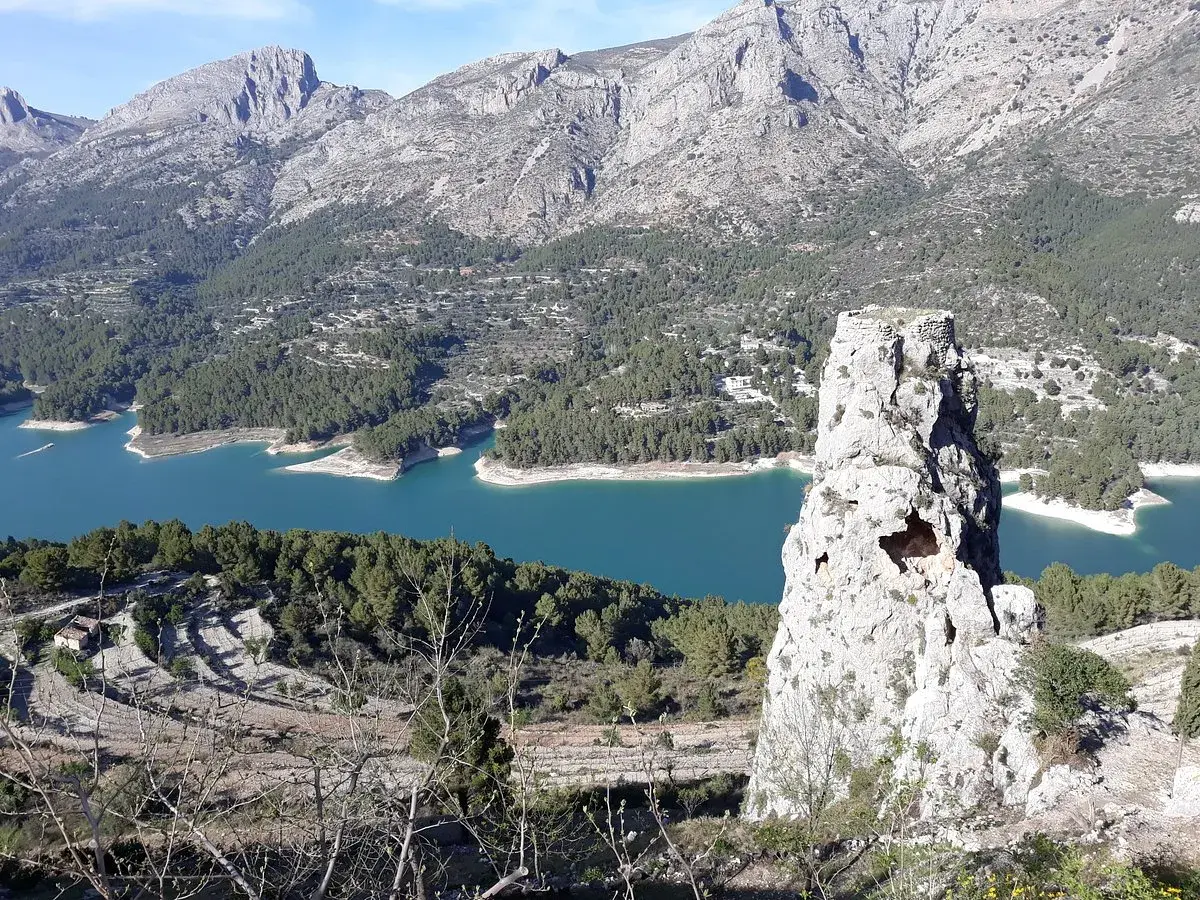 Impresionante vista del embalse de Guadalest con su agua turquesa, rodeado de montañas y pinos. Una roca solitaria se alza como centinela.